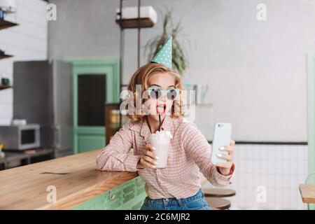 Beautiful lady in sunglasses and birthday cap sitting at the bar counter with milkshake and taking cute photos on her cellphone in cafe. Stock Photo