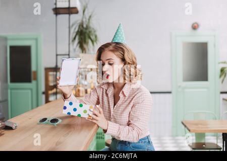 Young lady in birthday cap sitting at the bar counter and amazedly opening present box while spending time in cafe. Stock Photo