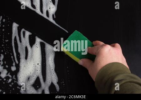 The texture of the soap foam stains marks on a black background. The concept of cleaning, and disinfection. and a hand with a green-yellow sponge wiping it off Stock Photo