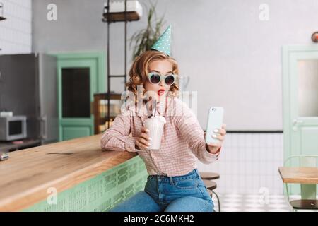 Young pretty lady in sunglasses and birthday cap sitting at the bar counter with milkshake and taking cute photos on her cellphone in cafe. Stock Photo