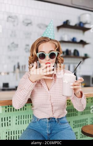 Young pretty lady in sunglasses and birthday cap sitting at the bar counter and drinking milkshake in cafe. Stock Photo