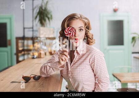 Pretty lady sitting at the bar counter and covering her eye with lollipop candy while amazedly looking in camera in cafe. Stock Photo