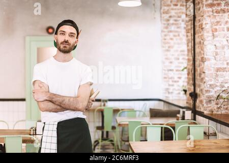 Young man in black cap,white t-shirt and apron standing and dreamily looking in camera while working in cafe. Stock Photo