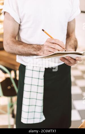 Close up photo of waiter standing with pencil and writing order in notebook in cafe. Stock Photo