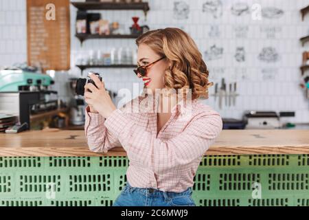Young smiling lady in sunglasses sitting at the bar counter and taking photos on her little camera while spending time in cafe. Stock Photo