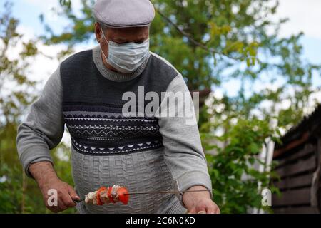 Mature man in medical mask impales raw shashlik on skewer. Older male holds skewer with barbecue. Concept of picnic outdoor. Stock Photo