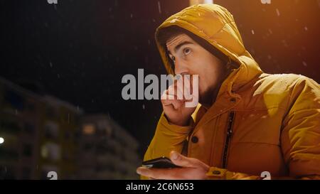 Young man in a hoodie messaging with the phone in his hand on a snowy night street Stock Photo