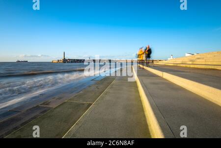 A couple walk on the steps at Margate seafront with the harbour arm in the background.
