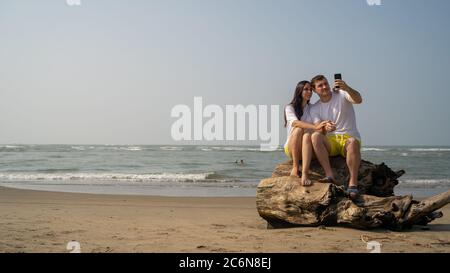 Happy couple sitting on driftwood near sea. Loving couple hugging and taking selfies while sitting on log during date on beach against waving sea and Stock Photo
