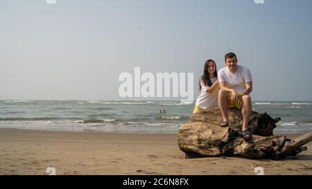 Happy couple posing on driftwood near sea. Loving couple hugging during date on beach against waving sea and cloudless sky. Stock Photo