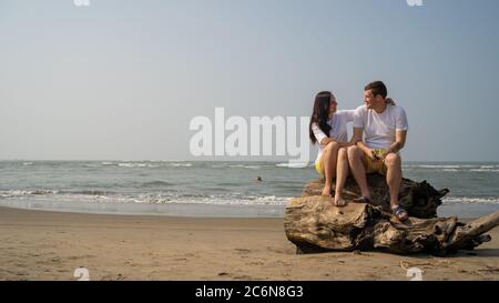 Happy couple sitting on driftwood near sea. Loving couple hugging and touching noses while sitting on driftwood during date on beach against waving Stock Photo