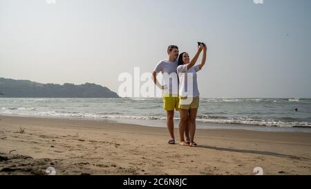 Happy couple taking selfies near sea. Loving couple embracing during date on beach against waving sea and cloudless sky. Stock Photo