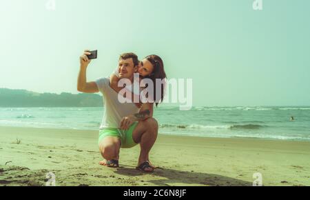 Happy couple taking selfies near sea. Loving couple embracing during date on beach against waving sea and cloudless sky. Stock Photo