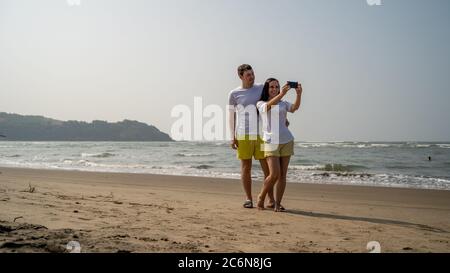 Happy couple taking selfies near sea. Loving couple embracing during date on beach against waving sea and cloudless sky. Stock Photo