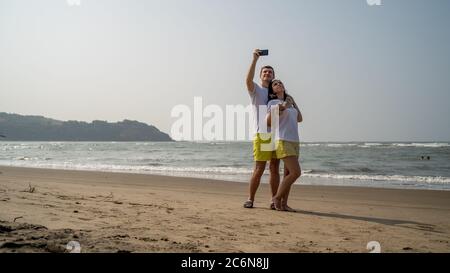Happy couple taking selfies near sea. Loving couple embracing during date on beach against waving sea and cloudless sky. Stock Photo