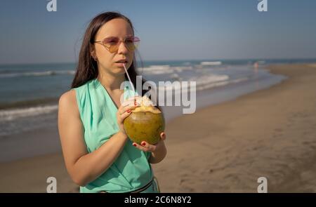 Young woman in sunglasses drinking coconut near sea or ocean. Female tourist enjoying tropical drink, sipping coconut water through straw on sandy Stock Photo