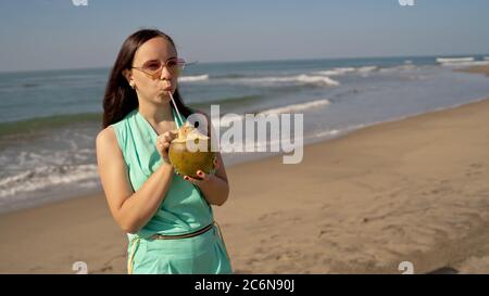 Young woman in sunglasses drinking coconut near sea or ocean. Female tourist enjoying tropical drink, sipping coconut water through straw on sandy Stock Photo