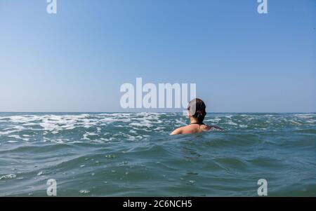 Young woman in black swimsuit spends time in sea. Adult lady swims towards the waves. Stock Photo