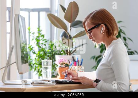 Freelancer woman taking notes with a pen, working on computer, using calculator to calculate the invoice, planning expenses from home surrounded by pl Stock Photo