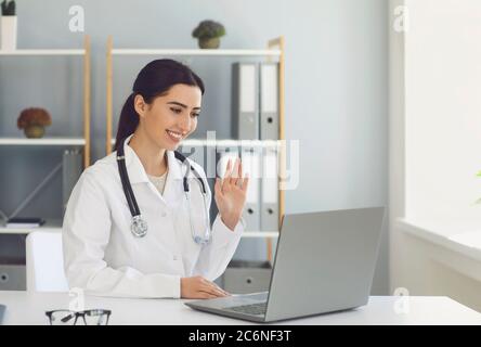 Hispanic female doctor waving to a patient has a video conference laptop sitting at a table in a clinic office. Stock Photo