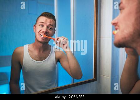 Morning hygiene routine. Man brushing teeth and looking in mirror of home bathroom. Stock Photo