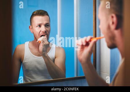 Morning hygiene routine. Man brushing teeth and looking in mirror of home bathroom. Stock Photo