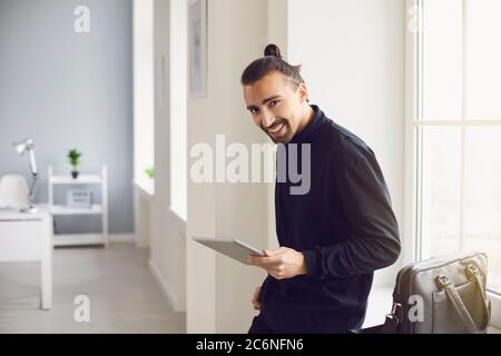 A positive guy smiling standing with a tablet in his hand in the office at the workplace. Stock Photo