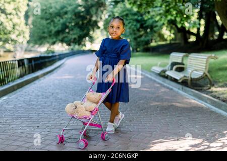 Little 5 years old African kid girl walk and playing with her toy stroller in park. Cute little dark skinned baby girl with a pram and teddy bear in Stock Photo