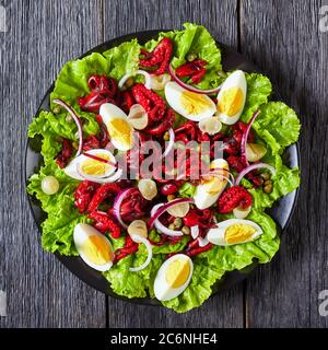 steamed baby octopus salad with hard-boiled eggs, capers, marinated onion on lettuce leaves on a black plate on a dark wooden table, horizontal view f Stock Photo