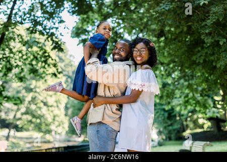 Happy joyful young African family, father, mother and little daughter, having fun outdoors, playing together in summer park. Mom, dad and kid laughing Stock Photo