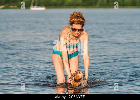 Attractive mature woman enjoying playing with her dog in whater during summer. Summer time and lifestyle. Dachshund swims in the river. An elderly Stock Photo