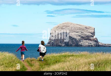 North Berwick, East Lothian, Scotland, United Kingdom, 11th July 2020. UK Weather: a very breezy sunny day in the seaside town along the shore of the Firth of Forth. The Bass Rock gannet colony is lit up in the sunshine as two young girls walk on the clifftop coastal path Stock Photo