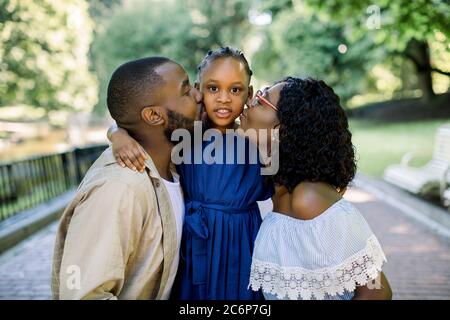 Adorable summer portrait of young african family of three posing together outdoors in beautiful park. Lovely parents are kissing their little daughter Stock Photo