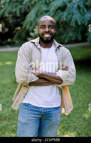 Portrait of young attractive bearded black man in casual shirt and jeans, standing at the park on a summer day, with happy smile and arms crossed Stock Photo
