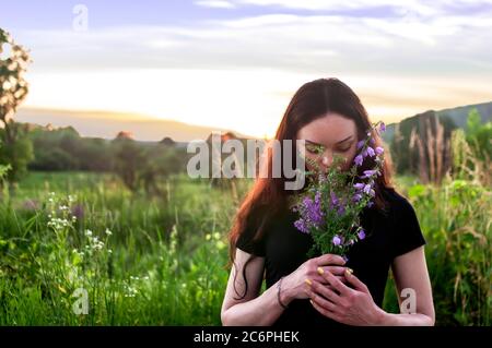 photo of a young girl in a black T-shirt on an evening summer walk with a bouquet of forest flowers on a background of sunset Stock Photo