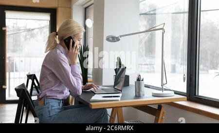 Young Caucasian female employee use laptop talking on cellphone Stock Photo