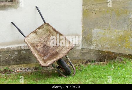 Old builders wheelbarrow resting against wall and standing on grass Stock Photo