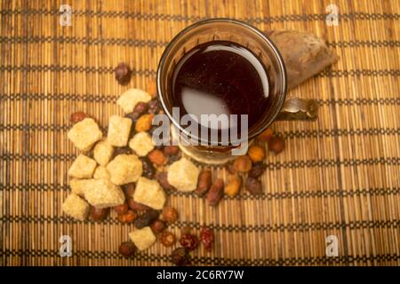 A cut-glass Cup of tea in a vintage Cup holder, dried rosehip fruit and chunks of brown cane sugar on a cane Mat. Close up Stock Photo