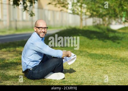 Side view portrait of smiling bald man sitting on green grass outdoors and looking at camera while enjoying sunlight, copy space Stock Photo