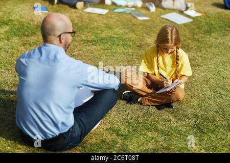 Full length portrait of blonde teenage girl sitting on green grass in sunlight and writing in notebook during outdoor class with teacher, copy space Stock Photo