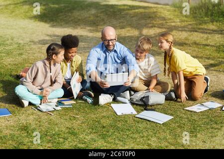 Full length portrait of smiling male teacher talking to group of children while sitting on green grass and enjoying outdoor class in sunlight, copy sp Stock Photo