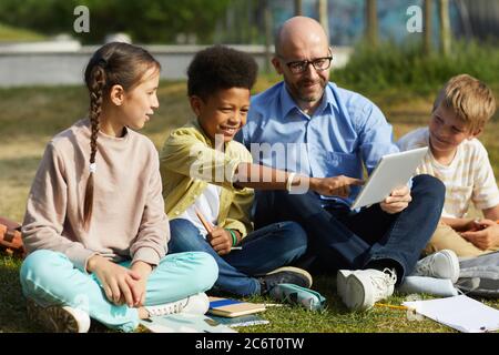 Full length portrait of male teacher talking to group of children while sitting on green grass and enjoying outdoor class in sunlight Stock Photo