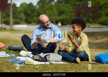 Full length portrait of smiling bald teacher talking to African-American boy holding model planet while enjoying outdoor astronomy lesson in sunlight, Stock Photo