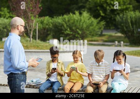 Multi-ethnic group of children using digital tablets while sitting in row on bench during outdoor lesson with smiling male teacher, copy space Stock Photo