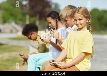 Multi-ethnic group of children using tablets and smartphones while sitting in row outdoors in sunlight, focus on smiling blonde girl in foreground, co Stock Photo