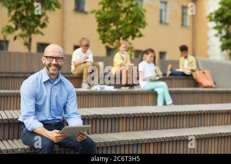 Portrait of smiling male teacher looking at camera while holding digital tablet sitting on bench outdoors with group of schoolchildren in background, Stock Photo