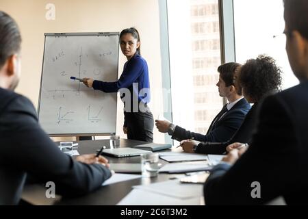 Serious young skilled female speaker pointing at charts on whiteboard. Stock Photo