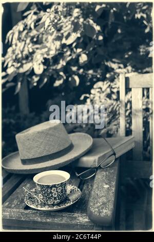 A gentleman's straw hat, cup of tea, reading book and glasses left on a table between a 'his' and 'hers' garden wooden seat on a summers afternoon. Stock Photo