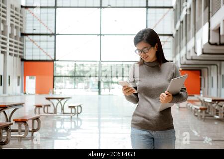Image of young woman student walking in the building using mobile phone holding laptop computer. Chats and correspondence, online communication. Stock Photo