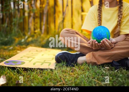 Close up of unrecognizable girl holding planet in hands while sitting on green grass and protesting for nature outdoors in sunlight, copy space Stock Photo